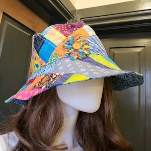 May include: A patchwork bucket hat with a colorful design. The hat features a variety of fabric squares in shades of blue, yellow, pink, and purple. A crocheted flower with a button center adorns the front. The brim is approximately 3 inches wide.