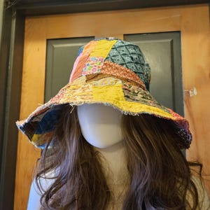 May include: A wide-brimmed bucket hat constructed from various fabric patches in shades of yellow, blue, pink, and orange. The hat features visible stitching throughout the patchwork design and has a frayed, raw edge along the entire brim circumference.