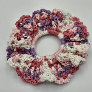 May include: A handmade scrunchie in shades of pink, purple, and white. The scrunchie is crocheted with a ruffled edge and a thick elastic band. The scrunchie is designed to hold hair in a ponytail or bun.