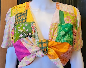 May include: A patchwork crop top with a bow tie in the front. The top is made of different fabrics in shades of green, yellow, pink, and orange. The bow tie is tied in a knot at the center of the chest.