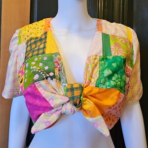 May include: A patchwork crop top with a bow tie in the front. The top is made of different fabrics in shades of green, yellow, pink, and orange. The bow tie is tied in a knot at the center of the chest.