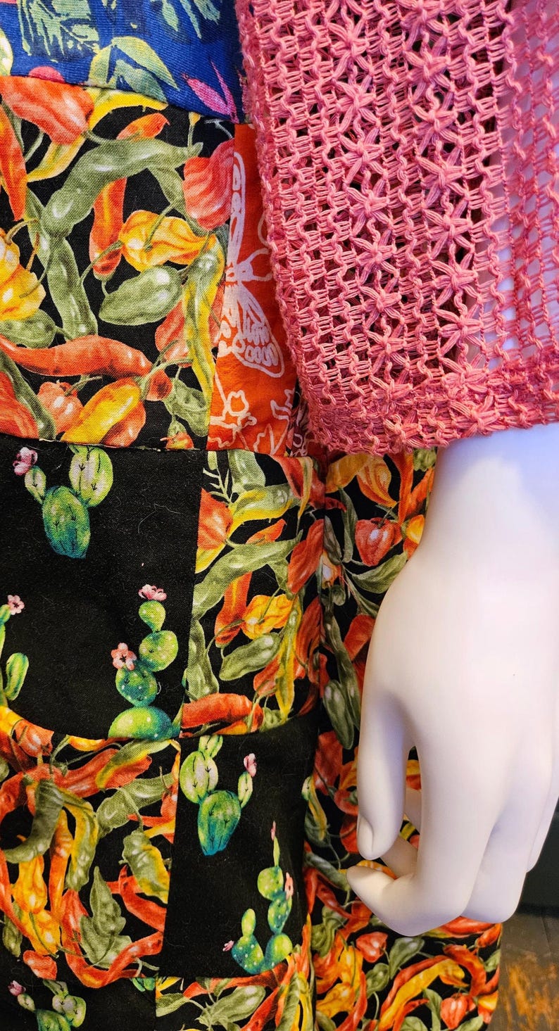 May include: A patchwork garment featuring a black fabric base with a vibrant print of orange and yellow chili peppers and green cactus plants, paired with a long-sleeved pink crocheted top with an open-weave texture.