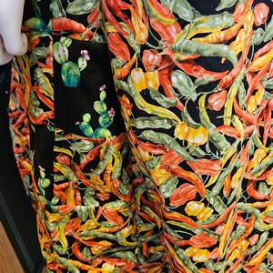 May include: A garment featuring a vibrant all-over print of red, orange, and yellow chili peppers mixed with green cactus plants on a black background. The fabric displays a dense pattern of various pepper shapes and small cacti with pink flowers.