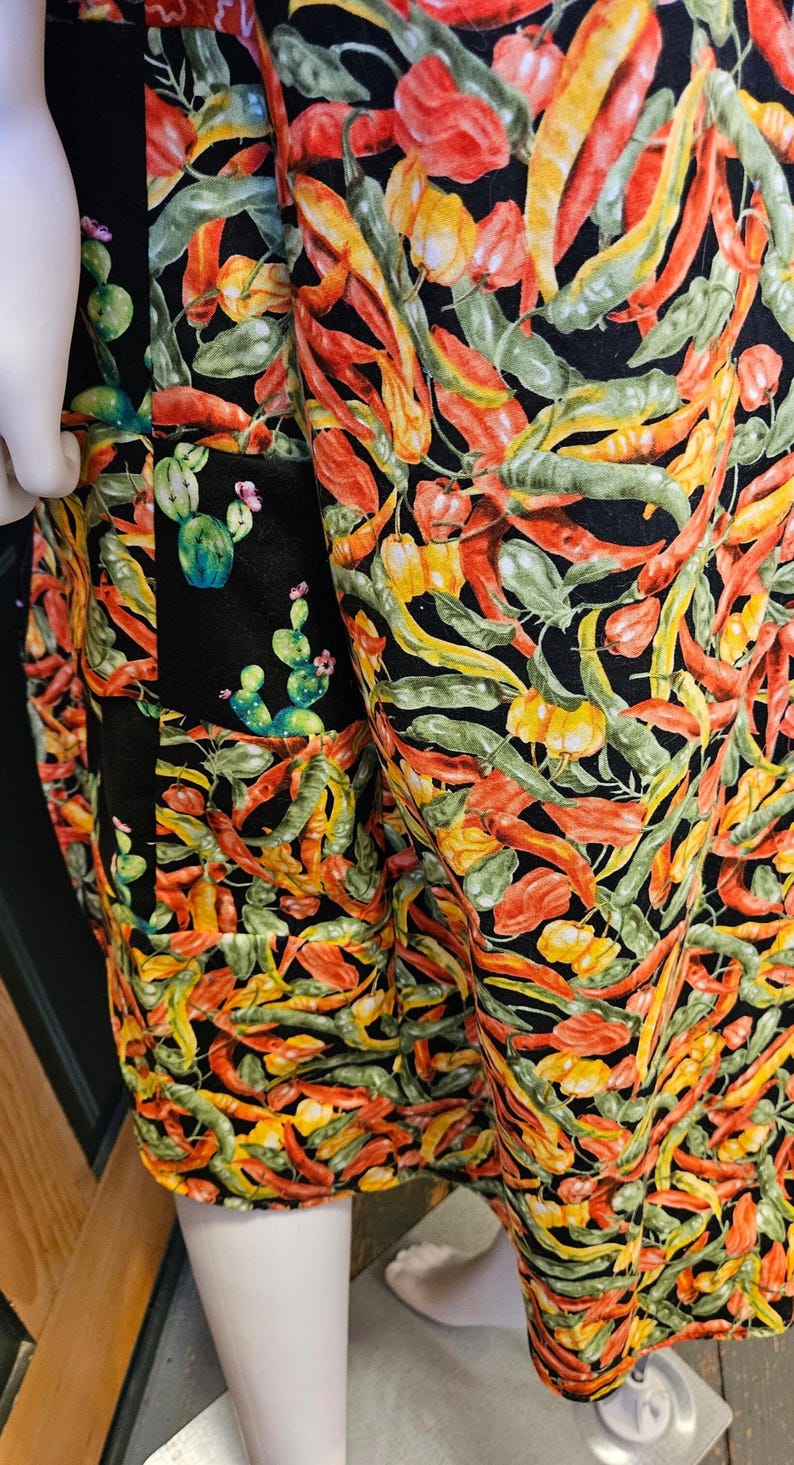 May include: A garment featuring a vibrant all-over print of red, orange, and yellow chili peppers mixed with green cactus plants on a black background. The fabric displays a dense pattern of various pepper shapes and small cacti with pink flowers.