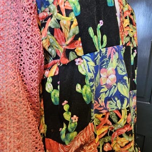 May include: A patchwork-style garment featuring a coral-colored open-knit sleeve and a main body composed of various fabric panels. The panels display vibrant prints of green cacti, pink flowers, and orange chili peppers on black and patterned backgrounds.