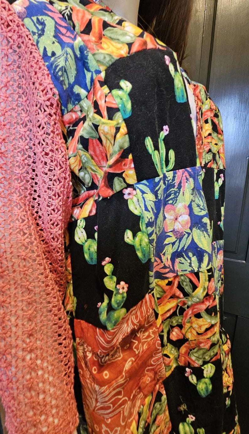 May include: A patchwork-style garment featuring a coral-colored open-knit sleeve and a main body composed of various fabric panels. The panels display vibrant prints of green cacti, pink flowers, and orange chili peppers on black and patterned backgrounds.