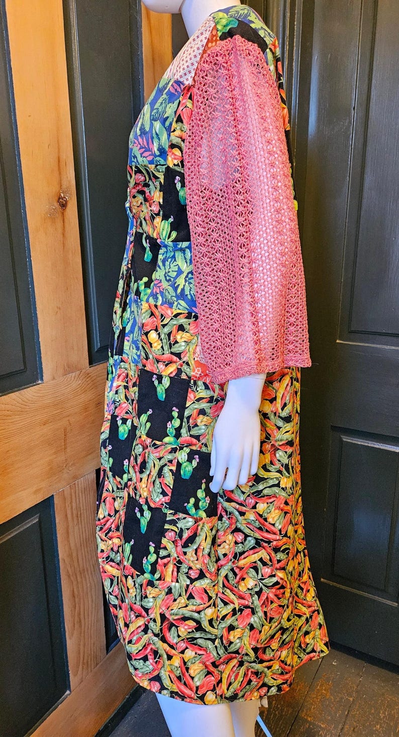 May include: A sleeveless dress featuring a patchwork design with black squares and vibrant, multi-colored botanical prints of cacti and chili peppers, complemented by a separate, textured, dusty pink, open-knit, three-quarter sleeve bolero.