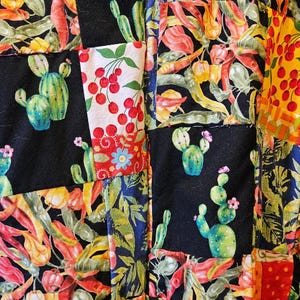 May include: A patchwork fabric featuring a variety of square panels with colorful prints. The patterns include green cacti, red cherries, yellow and red chili peppers, and lush green tropical leaves set against black and white backgrounds.