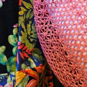 May include: A garment featuring a black base fabric with a vibrant, multi-colored botanical print of green cacti, orange peppers, and pink flowers, paired with a textured, open-weave coral-colored crochet overlay piece.