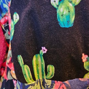 May include: A black fabric garment featuring a repeating pattern of green cacti with small pink flowers, interspersed with vibrant red and orange chili peppers and green foliage. The textile displays a dense, colorful botanical print.