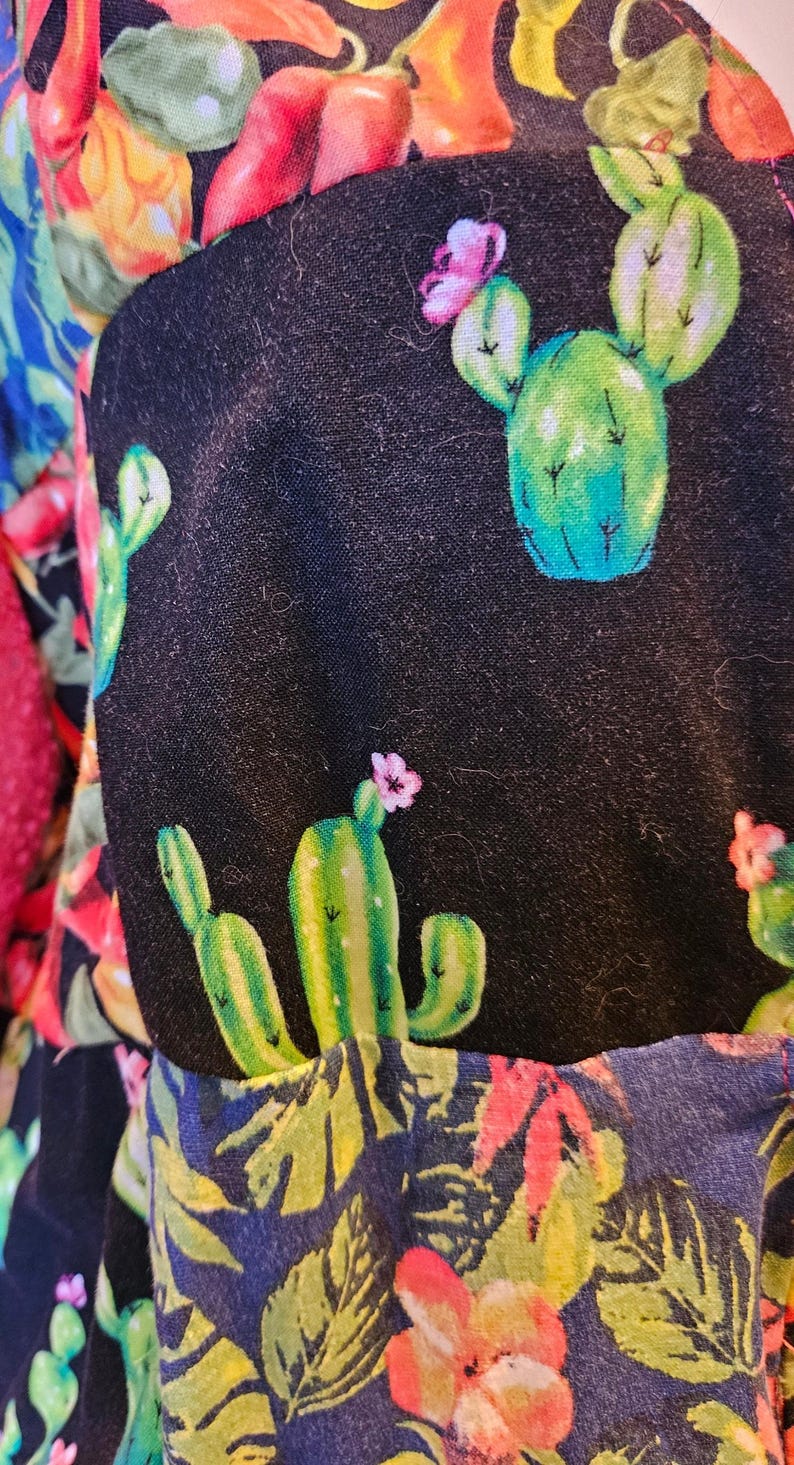 May include: A black fabric garment featuring a repeating pattern of green cacti with small pink flowers, interspersed with vibrant red and orange chili peppers and green foliage. The textile displays a dense, colorful botanical print.