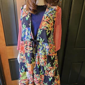 May include: A patchwork duster coat featuring a black fabric with a colorful cactus print, long coral-colored crochet sleeves, and a black drawstring waist. The outfit includes a matching patchwork bucket hat and patchwork trousers with fringe trim at the hem.