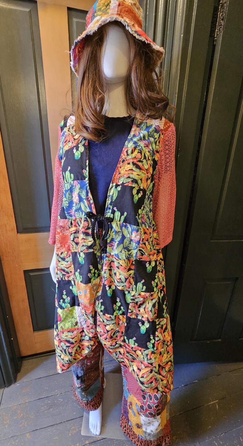 May include: A patchwork duster coat featuring a black fabric with a colorful cactus print, long coral-colored crochet sleeves, and a black drawstring waist. The outfit includes a matching patchwork bucket hat and patchwork trousers with fringe trim at the hem.
