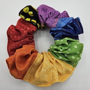 May include: A circular fabric hair scrunchie constructed from multiple segments of patterned cotton fabric in a rainbow spectrum, including black, purple, blue, green, yellow, orange, and red sections with visible zigzag stitching.