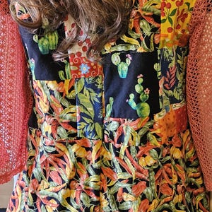 May include: A sleeveless dress featuring a patchwork design with black panels displaying green cacti and colorful sections of red chili peppers, yellow peppers, and cherries, paired with orange open-knit sleeves.
