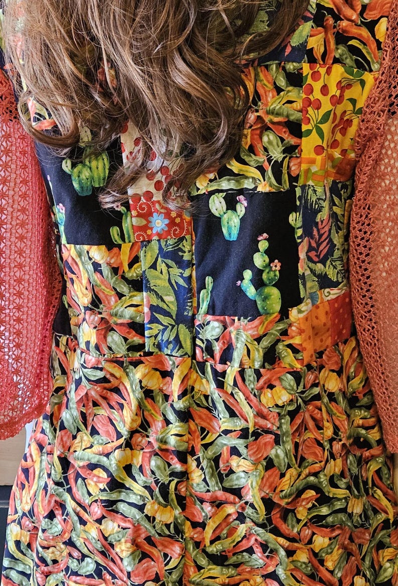 May include: A sleeveless dress featuring a patchwork design with black panels displaying green cacti and colorful sections of red chili peppers, yellow peppers, and cherries, paired with orange open-knit sleeves.