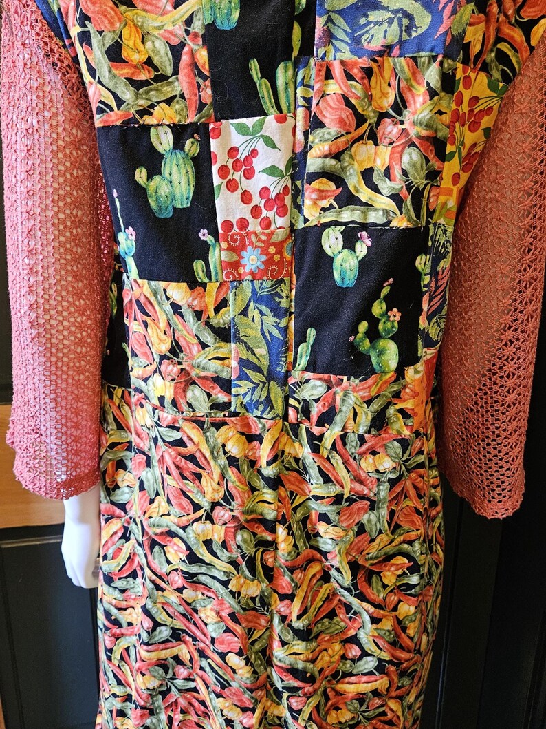 May include: A garment featuring a patchwork design of black fabric squares printed with green cacti and red cherries, interspersed with sections of vibrant chili pepper patterns. The sleeves are made of a textured, coral-colored mesh fabric.