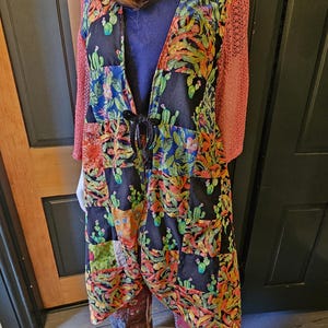 May include: A long, open-front duster cardigan featuring a patchwork design of black fabric printed with colorful cacti and succulents. The garment has three-quarter length sleeves made of a textured, coral-colored knit fabric and a black tie closure.