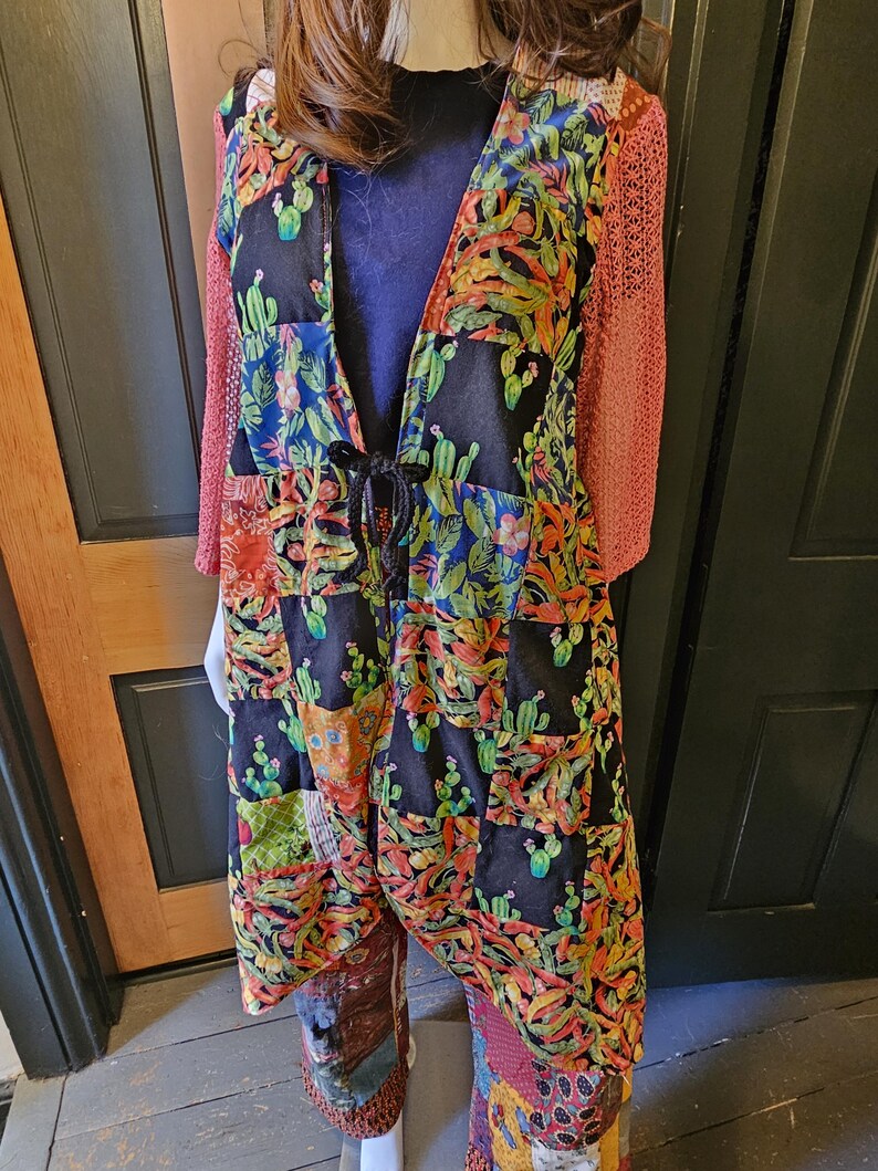 May include: A long, open-front duster cardigan featuring a patchwork design of black fabric printed with colorful cacti and succulents. The garment has three-quarter length sleeves made of a textured, coral-colored knit fabric and a black tie closure.