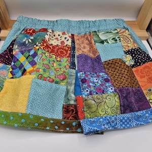 May include: A colorful patchwork skirt with a light blue elastic waistband. The skirt features a variety of patterned fabrics in shades of green, blue, yellow, orange, and purple. The hem is trimmed with a green fabric with blue polka dots.