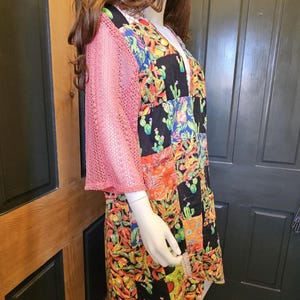 May include: A patchwork dress featuring a vibrant cactus and chili pepper print on a black background, paired with pink mesh sleeves and a matching multicolored patchwork bucket hat, displayed on a mannequin.