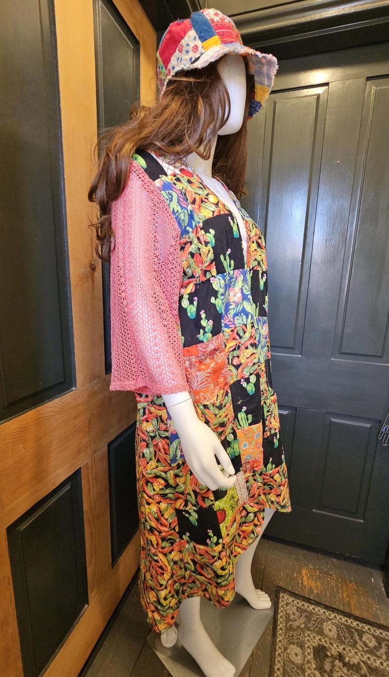 May include: A patchwork dress featuring a vibrant cactus and chili pepper print on a black background, paired with pink mesh sleeves and a matching multicolored patchwork bucket hat, displayed on a mannequin.