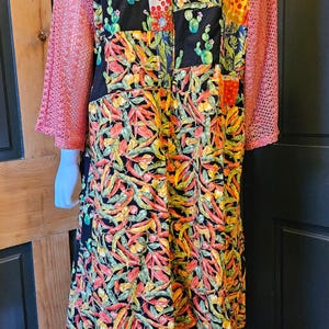 May include: A long-sleeved garment featuring a patchwork design with a black background, vibrant chili pepper prints, and cactus motifs. The sleeves are made of a textured, coral-colored open-knit fabric, contrasting with the colorful, busy pattern of the main body.