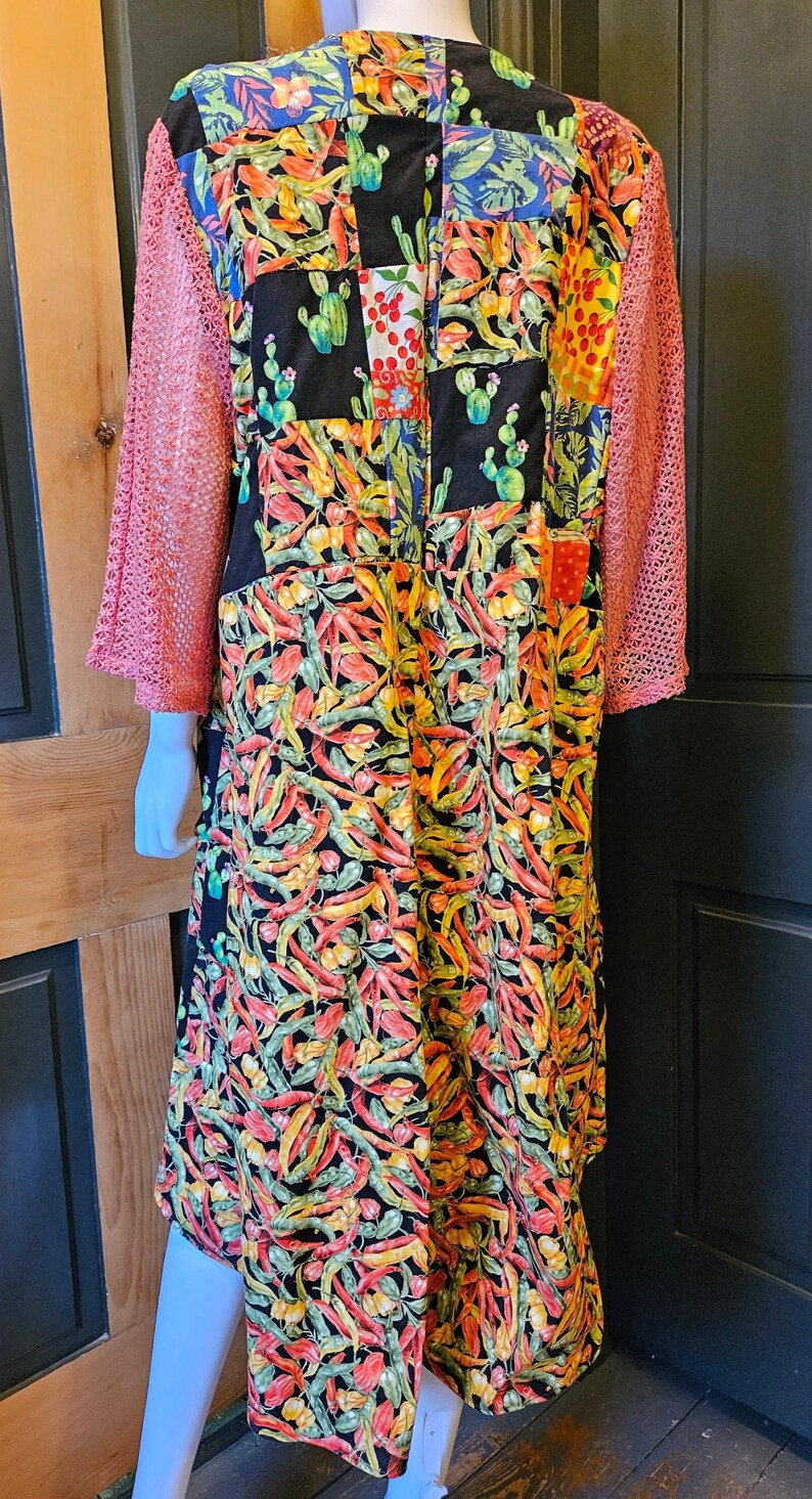 May include: A long-sleeved garment featuring a patchwork design with a black background, vibrant chili pepper prints, and cactus motifs. The sleeves are made of a textured, coral-colored open-knit fabric, contrasting with the colorful, busy pattern of the main body.
