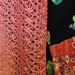 May include: A coral-colored crochet garment with an intricate openwork pattern featuring star-like motifs. Beneath the crochet layer, a black fabric displays a green cactus print with pink flowers, alongside a section of orange fabric.