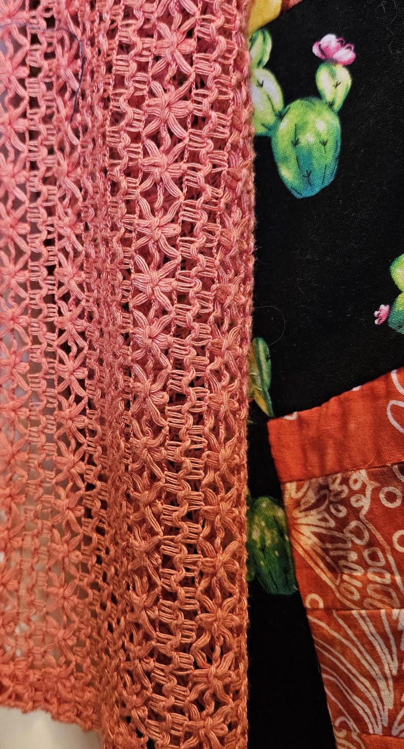 May include: A coral-colored crochet garment with an intricate openwork pattern featuring star-like motifs. Beneath the crochet layer, a black fabric displays a green cactus print with pink flowers, alongside a section of orange fabric.