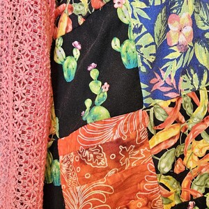 May include: A garment featuring a patchwork design with black fabric panels printed with green cacti and pink flowers, alongside panels of orange fabric with white leaf patterns and blue fabric with tropical foliage, paired with a coral crochet sleeve.