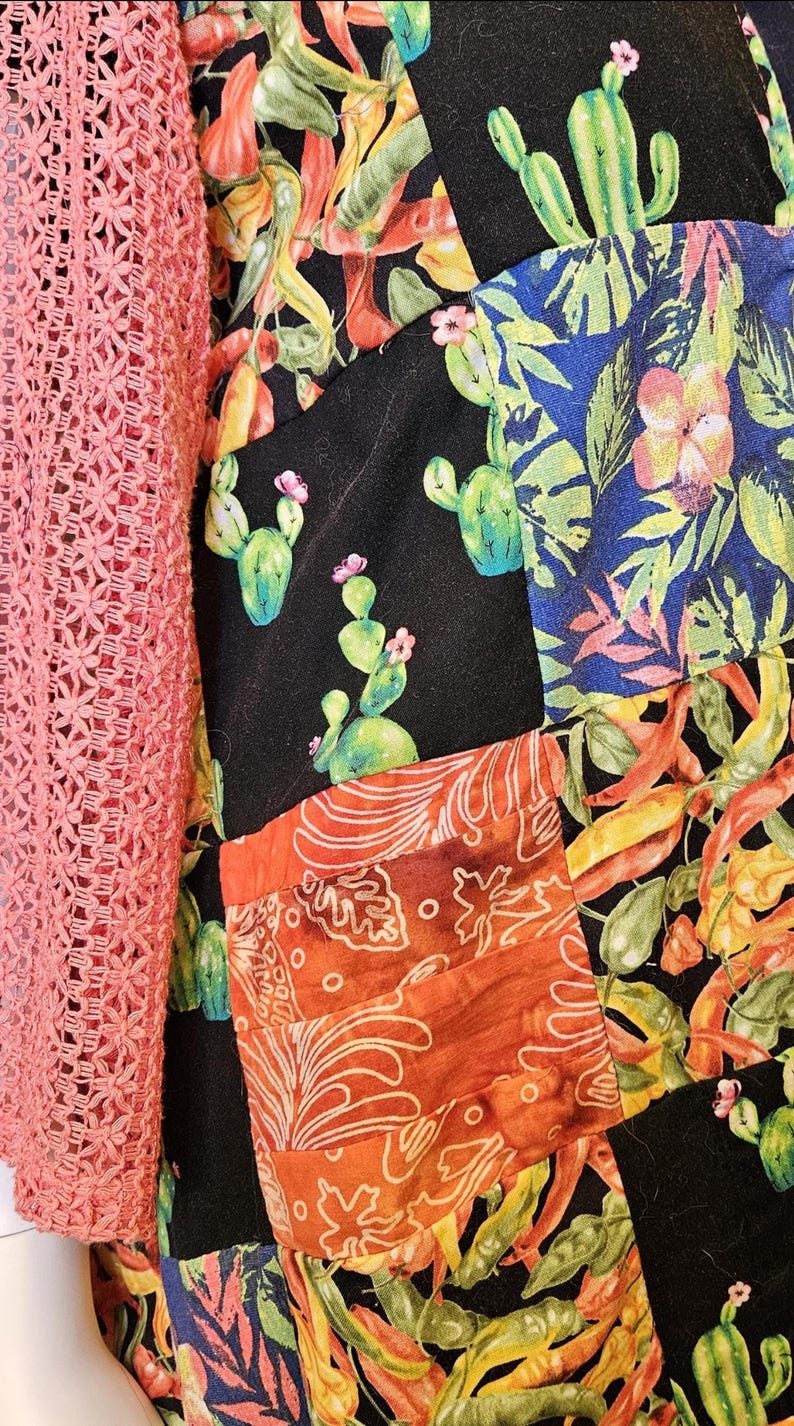 May include: A garment featuring a patchwork design with black fabric panels printed with green cacti and pink flowers, alongside panels of orange fabric with white leaf patterns and blue fabric with tropical foliage, paired with a coral crochet sleeve.
