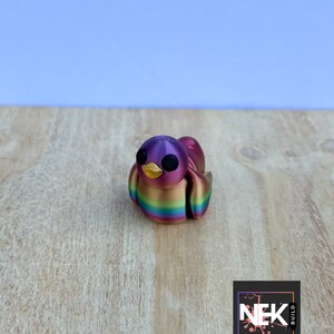 Op de afbeelding: Een klein, 3D-geprint vogelbeeldje met een regenboogkleurige afwerking. De vogel heeft zwarte ogen en een gele snavel. Het beeldje staat op een houten oppervlak.