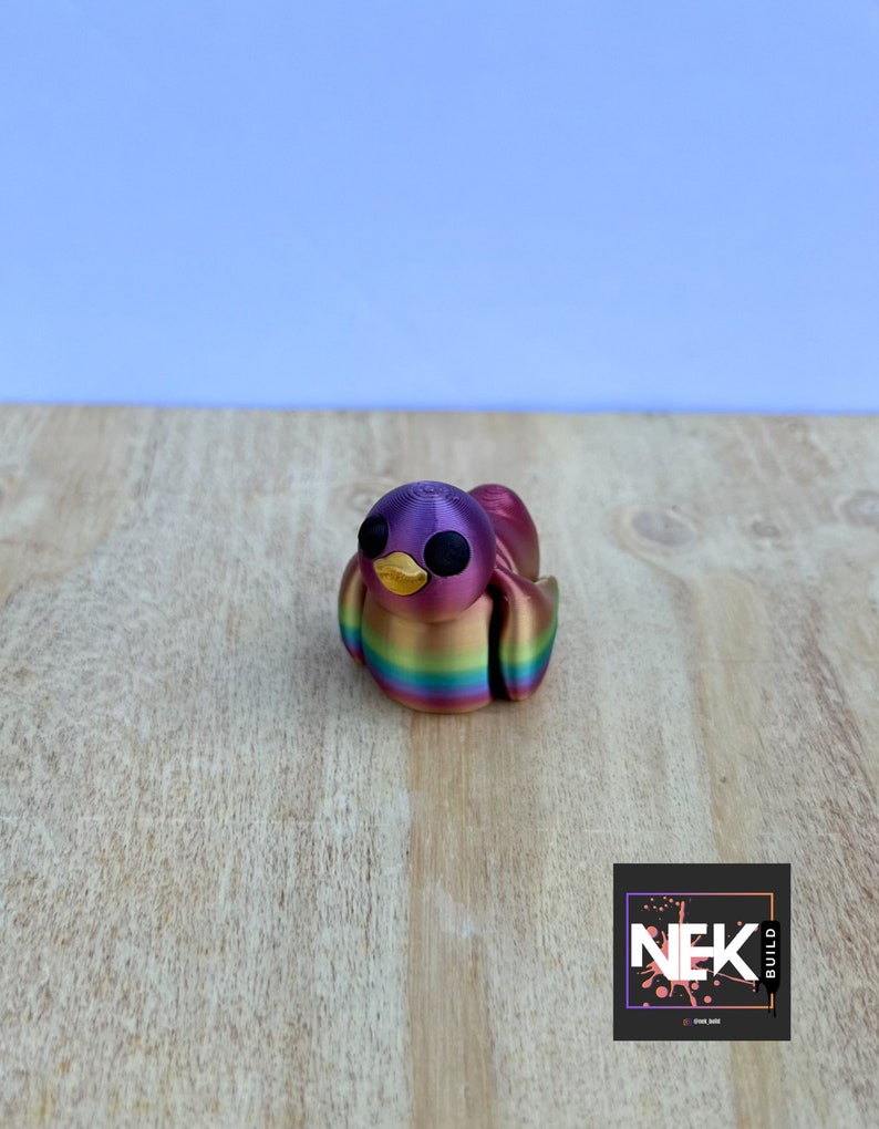 Op de afbeelding: Een klein, 3D-geprint vogelbeeldje met een regenboogkleurige afwerking. De vogel heeft zwarte ogen en een gele snavel. Het beeldje staat op een houten oppervlak.