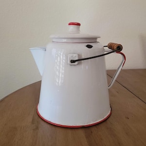 May include: White enamel camping coffee pot with a red rim and a wooden handle. The pot has a black metal handle and a red knob on the lid.