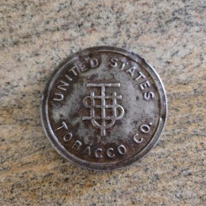Pode incluir: Uma ficha metálica circular e antiga com o texto "UNITED STATES TOBACCO CO." ao redor da borda. O centro apresenta um design estilizado "T" e "S". A ficha tem uma aparência desgastada e envelhecida.