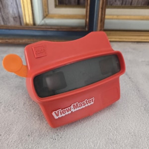 Könnte beinhalten: Ein rotes Plastik-View-Master-Spielzeug mit einem orangefarbenen Griff. Das Spielzeug hat zwei Sichtlinsen und die Worte "View-Master" sind in Weiß auf der Vorderseite aufgedruckt.