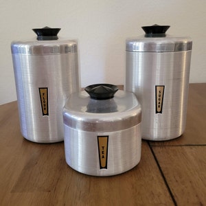 May include: Set of three vintage silver canisters with black lids and gold labels. The labels read "Flour", "Sugar", and "Tea".