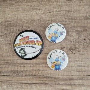 May include: Two round buttons featuring a blue and yellow cartoon bug design. The text on the buttons reads "Catch The Bug" and includes the year 2000. A third button is black with white text that reads "The Great Wal-Mart Film Round Up" and "Rediscover the Memories".