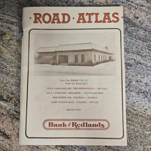 May include: A vintage "Road Atlas" booklet with a sepia-toned cover. The cover features the text "ROAD ATLAS" in brown letters and an image of a building. Below the image, the text reads "You Can BANK ON US!" and lists bank locations.