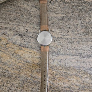 May include: A vintage wristwatch with a silver-colored back and a light brown leather strap. The watch has a round face and a buckle closure. The strap is approximately 9 inches long.