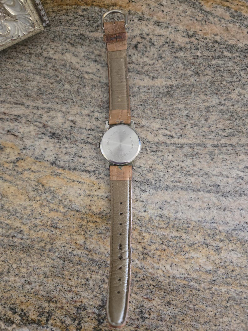 May include: A vintage wristwatch with a silver-colored back and a light brown leather strap. The watch has a round face and a buckle closure. The strap is approximately 9 inches long.