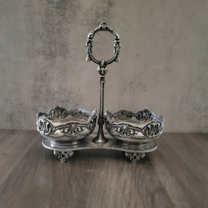 Op de afbeelding: Zilveren serveerschaal met twee kommen, elk met een versierd ontwerp. De schaal heeft een handvat met een decoratieve ring.