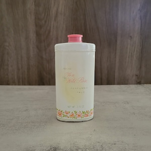 Pode incluir: Uma lata de metal branca com tampa rosa contendo talco perfumado Avon To a Wild Rose. A lata tem um design floral com rosas cor-de-rosa e folhas verdes. O texto na lata diz "Avon To a Wild Rose Perfumed Talc" e "Net Wt. 2.75 oz."