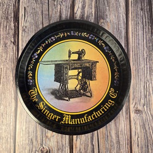 May include: A round black metal tray featuring a vintage Singer sewing machine illustration. The tray has a colorful gradient background and the text "The Singer Manufacturing Co." in gold lettering around the edge. Decorative floral accents are also present.