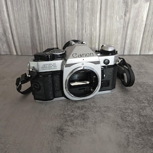 May include: Silver and black Canon AE-1 Program 35mm film camera with a black strap. The camera body has the Canon logo and AE-1 Program printed on it. The camera is on a grey surface.