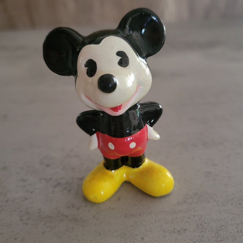 Op de afbeelding: Een klein keramiek beeldje van Mickey Mouse, gemaakt in zwart en wit. Hij draagt een rode broek met witte stippen en gele schoenen. Hij staat met zijn armen gekruist en heeft een vriendelijke glimlach.
