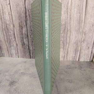 May include: A green hardcover book with the title "Babar Comes to America" by  "De Brunhoff" on the spine. The cover has a repeating pattern of small brown and white dots.