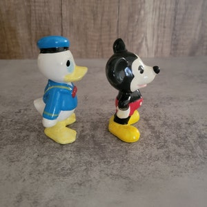 Op de afbeelding: Een keramiek beeldje van Donald Duck met een blauwe matrozenpet en een blauw-wit gestreept matrozenoverhemd.  Naast hem staat een keramiek beeldje van Mickey Mouse met een rode broek en zwarte schoenen.