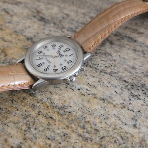 May include: A silver-toned wristwatch with a white face and black numerals. The watch has a light brown leather band. The watch face reads "TIME QUARTZ".